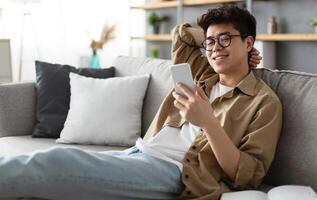 Enjoying Weekend. Handsome Asian Man In Glasses Using Smartphone While Sitting Leaning On Couch At Home, Happy Guy Relaxing In Living Room, Browsing Internet Or Messaging With Friends, Selective Focus photo