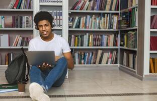 Young african american student with headset sitting on the floor, leaning on bookshelf and using laptop at library, free space photo