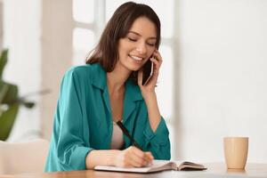 Note Taking. Portrait of female student making phone call and writing memo in her diary, copyspace photo