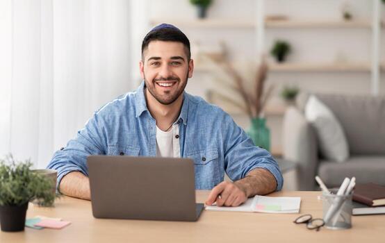 hogar oficina concepto. sonriente judío hombre vistiendo kipa sentado a escritorio, trabajando en ordenador personal ordenador portátil adentro en vivo habitación. contento persona de libre dedicación posando y mirando a cámara. distante trabajo, Internet y tecnología foto