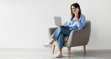 Full length of Caucasian lady using laptop and speaking on smartphone while sitting in armchair against white studio wall, free space. Lovely young woman having phone conversation, working on pc photo