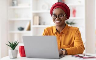 Cheerful millennial black woman entrepreneur working from home, wearing red headscarf and eyeglasses, sitting at workdesk and typing on laptop keyboard, chatting with clients, copy space photo