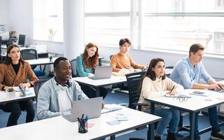 Lecture Concept. Group of interested multicultural mixed race group of students sitting at tables with pc in modern classroom, listening to teacher, taking notes writing in notebooks, back to study photo