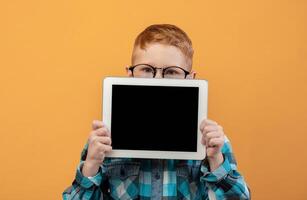 Ginger boy in eyeglasses hiding behind digital tablet with empty screen over yellow studio background. Cute redhead schoolboy showing pad, mockup, advertisement for cute application photo