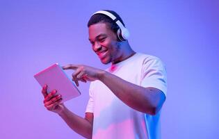 Interesting App. Portrait Of Excited African Guy In Wireless Headphones Using Digital Tablet, Millennial Black Man Playing Games Or Browsing Internet While Standing Under Purple Neon Light photo