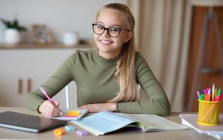 Cheerful teenager doing homework, making notes or writing essay. Pretty blonde school girl in glasses writing in notepad and smiling at camera, enjoying studying, home interior, copy space photo