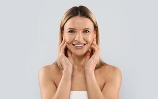 Face injections concept. Pretty blonde middle aged woman wrapped in white bath towel touching her face skin and smiling, enjoying results of face care treatment, grey studio background photo