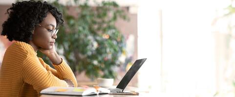 Pensive afro woman editor reading text on laptop, taking notes, working at cafe, copy space photo