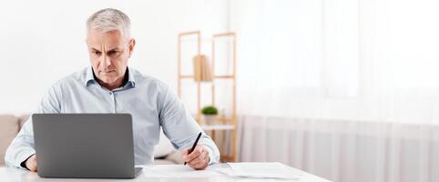 Budget Planning. Pensive Senior Man Using Laptop And Writing In Notebook Sitting At Desk At Home, Empty Space photo