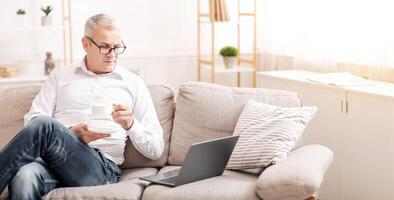 In The Morning. Senior Man Using Laptop Computer Having Coffee Sitting On The Couch At Home, Copyspace photo