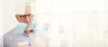 Business Strategy. Pensive Senior Manager In Specs Looking At Sticky Notes On Window At Office, Copyspace photo