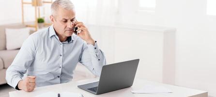 Business Calls Concept. Concerned Senior Man Talking On Mobile Phone Sitting At Desk, Looking At Laptop, Empty Space photo