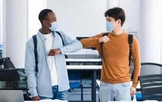 Elbow Bump. Smiling multicultural students wearing medical face masks greeting each other at classroom. Asian and african american men avoid touching, studying in university, standing at lecture hall photo