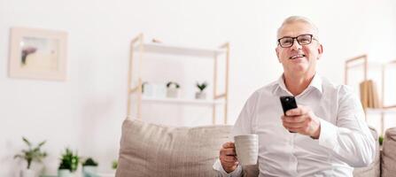 Watching Movie On Tv. Smiling Senior Man Sitting On Couch With Remote Controller, Holding Coffee Mug, Copyspace photo