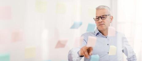 Project Planning. Serious Mature Businessman Reading Colorful Sticky Notes On Glass Visualisation Board In Office photo