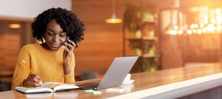 Black business woman taking notes while talking with client on smartphone, working with laptop at cafe, copy space photo