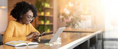 Happy black girl using mobile phone, working with laptop in cafe, side view, copy space photo