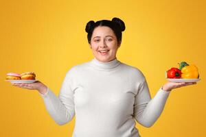 Smiling Plus Sized Girl Holding Plates With Fresh Vegetables And Donuts In Both Hands, Posing Over Yellow Background, Panorama photo