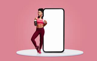A fit woman in workout clothing stands happily with a drink in hand beside a large smartphone. The background features a soft pink color, highlighting her cheerful demeanor and fitness lifestyle. photo