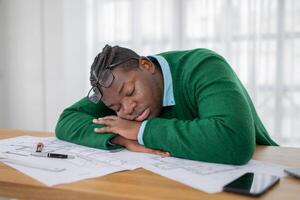 A young black man in a green sweater rests his head on his arms at a desk cluttered with design drafts. Warm sunlight fills the modern office where he appears to be exhausted from work. photo
