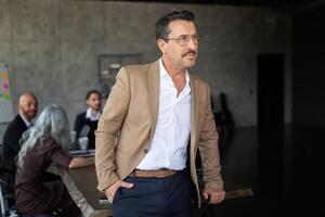 A man in a beige blazer stands confidently while engaging in a business meeting. Behind him, a diverse group collaborates at a large table, sharing thoughts in a stylish, contemporary office. photo