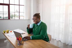 A cheerful black man in a green sweater sits at a wooden desk, sipping coffee from a mug while working on his laptop. The sun streams through the windows, creating a warm atmosphere. photo