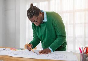 A focused black guy architect is diligently sketching blueprints on a large table in a modern office. Natural light streams in through the windows, illuminating the workspace and materials. photo