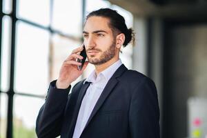 A business professional wearing a suit is talking on a mobile phone inside a modern office setting. Sunlight streams through large windows, creating a dynamic atmosphere. photo