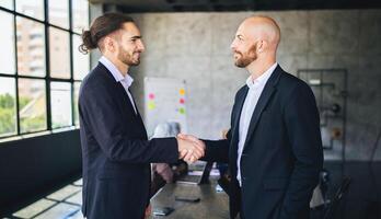 Two men in suits greet each other with a handshake in a contemporary office. A group of colleagues is engaged in discussion in the background, emphasizing collaboration and professionalism. photo