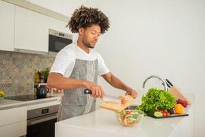 A black man with curly hair is chopping vegetables on a wooden cutting board in a stylish kitchen. Fresh ingredients are neatly arranged, showcasing a healthy cooking environment. photo