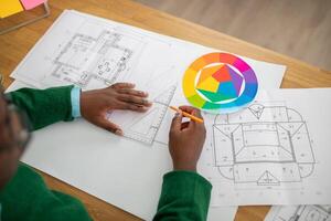 A person examines detailed architectural plans while using a color palette and drawing tools. The creative workspace is filled with sketches and planning materials, reflecting an artistic process. photo