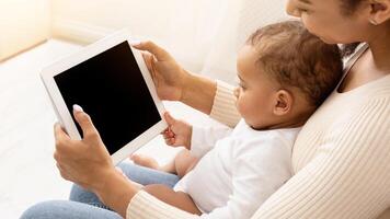 African American Mom With Toddler Baby Using Digital Tablet With Blank Screen Sitting On Sofa At Home. Side View Of Mom And Infant Using Computer Together. Application Advertisement. Mockup photo