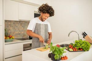 A black man in an apron joyfully slices cucumbers on a cutting board in a bright, modern kitchen filled with fresh vegetables and vibrant greens, preparing a nutritious meal. photo