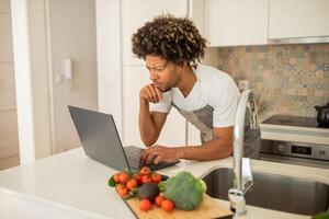 A black man with curly hair is focused on his laptop while surrounded by fresh vegetables in a stylish kitchen. He balances cooking and work, showcasing modern lifestyle choices. photo