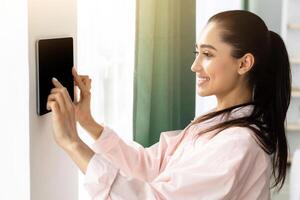Innovative Technology. Millennial Woman Using Digital Tablet With Empty Screen On Wall With Smart Home Control System App Adjusting House Automation Processes And Settings Standing Indoors. Mockup photo