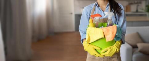 The individual in gloves carries a vibrant green container filled with cleaning tools and supplies while preparing to clean a well-lit living space with soft furnishings. photo