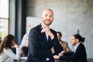 A young man with a beard confidently poses in a stylish suit at a meeting table while colleagues engage in discussion. The modern office has large windows and a contemporary design. photo