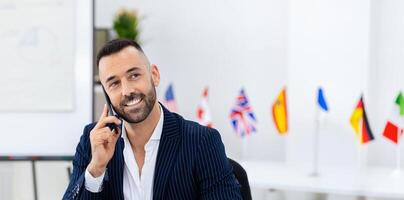 A well-dressed man is engaged in a phone conversation, smiling as he communicates. Flags from various nations are displayed in the background, indicating an international environment. photo