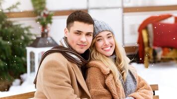 A young couple sits closely on a bench in a snowy park, smiling at the camera. They wear warm clothing and the atmosphere is cheerful with light snow falling around them. photo