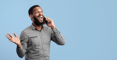 Unbelievable News. Portrait of surprised shocked black guy talking on his cellphone standing isolated over blue studio background with copy space, banner. Astonished man gesturing and speaking photo