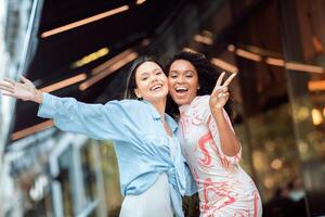 Two women are enjoying a sunny day outside a stylish cafe, smiling and striking a playful pose. They radiate happiness as they celebrate their time together. photo