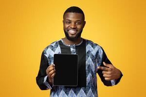 Smiling black guy pointing at digital tablet with empty screen, wearing traditional african costume. Positive african american young man showing newest application over pink studio background photo