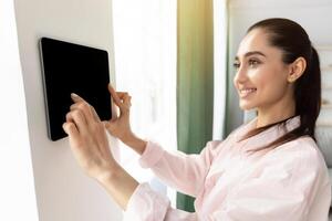 Innovative Technology For Apartment. Smiling Lady Using Digital Tablet On Wall With Smart Home Control System App Adjusting House Automation Settings, Standing In Living Room, Selective Focus photo