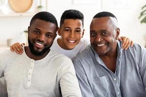 Generation. Smiling Black Boy Embracing His Dad And Grandfather Posing To Camera At Home, Sitting Together On Couch In Living Room. photo