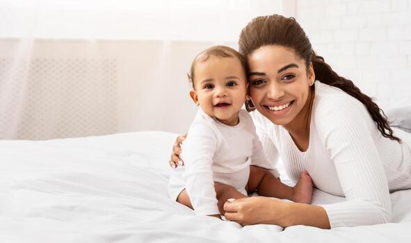 contento maternidad. africano madre abrazando bebé niñito posando sentado en cama en dormitorio a hogar, sonriente a cámara. alegría de siendo mamá, maternidad salir y niño cuidado concepto foto