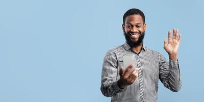 Hello. Front view portrait of excited black guy having call and waving hand at camera, using cell phone, copyspace photo