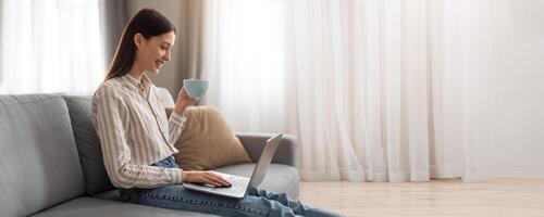 A young woman sits comfortably on a couch, sipping coffee from a light blue cup while focused on her laptop. Sunlight streams in through sheer curtains, creating a cozy atmosphere. photo