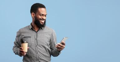 Communication Concept. Happy african american guy watching , chatting on smartphone standing over blue studio background, drinking takeaway coffee from cup. Man browsing mobile internet photo