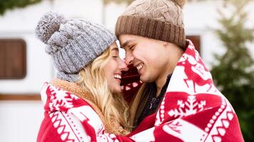 A young couple smiles at each other while wrapped in a red blanket, wearing cozy hats. They enjoy a cold winter morning surrounded by trees, radiating warmth and affection. photo