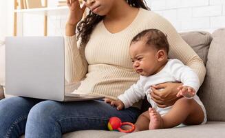 Exhausted African American Mother Hugging Crying Baby Using Laptop Browsing Internet Sitting At Home. Mom Trying To Work Online On Computer While Infant Son Screaming Feeling Bad photo